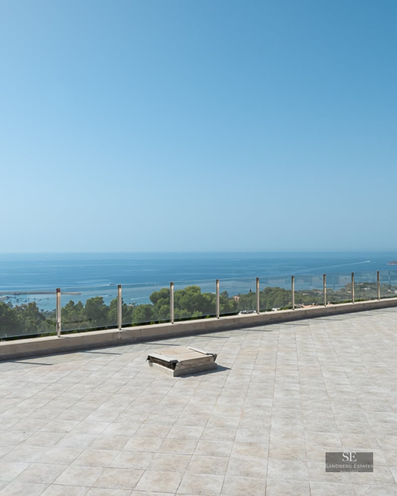 A large stone-tiled terrace with a glass railing overlooking a vast blue ocean under a clear sky.