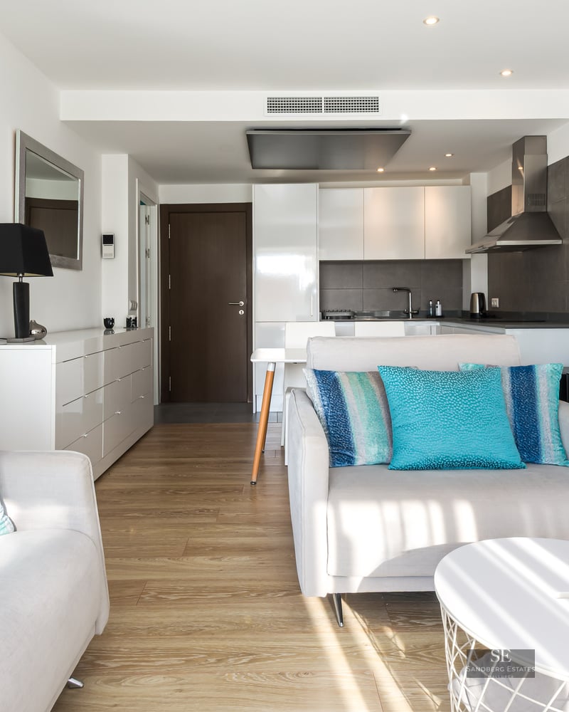 Salon lumineux avec canapés blancs, coussins turquoise, parquet en bois et cuisine moderne en arrière-plan.