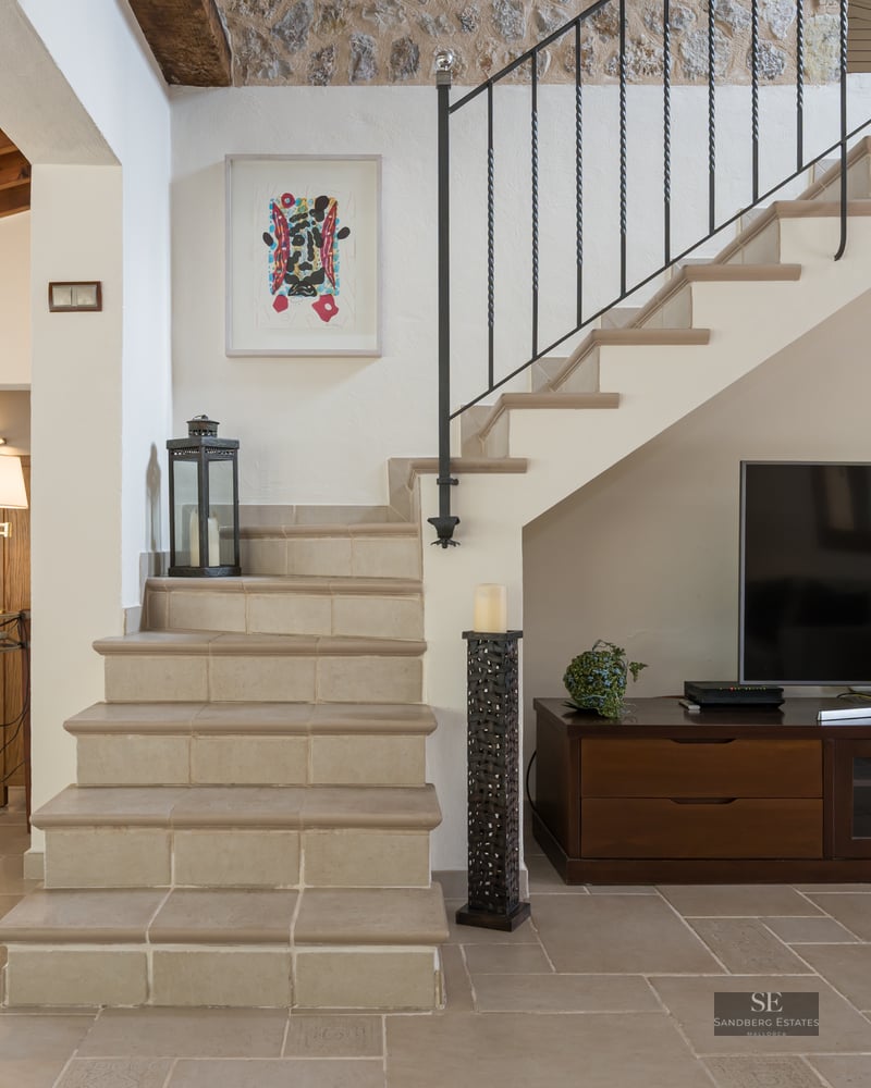 Interior view featuring a stone staircase, wooden TV console, and dining area with exposed ceiling beams.