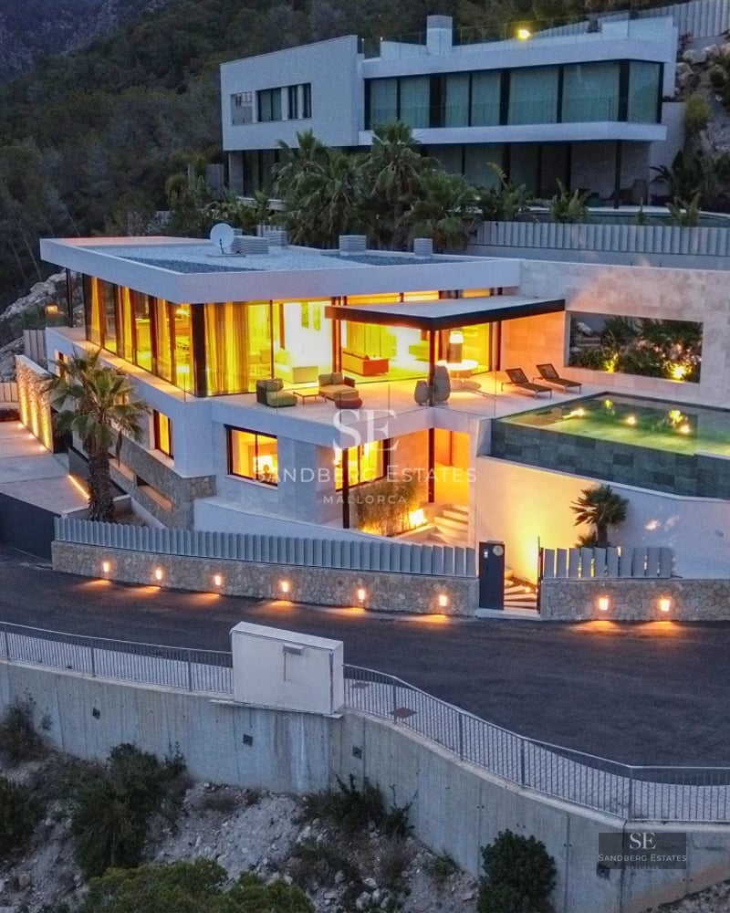 View of a luxurious villa with infinity pool, landscaped gardens, and modern architecture. Stone facade and large windows.