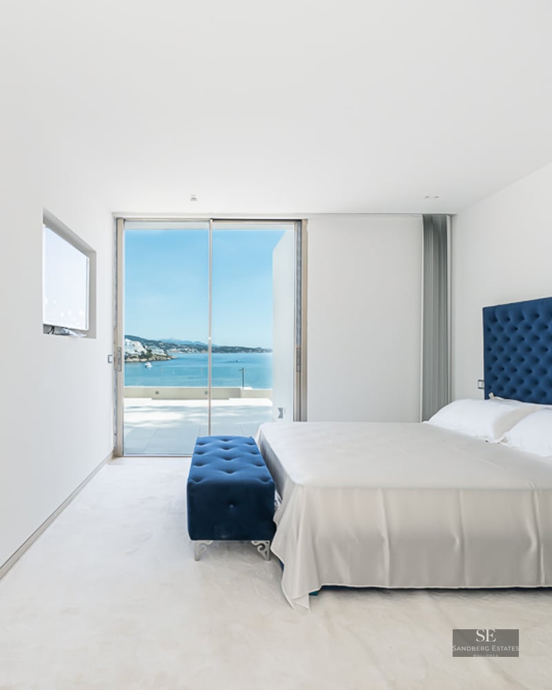 Modern bedroom with blue tufted headboard, white linens, and large glass doors overlooking the ocean.