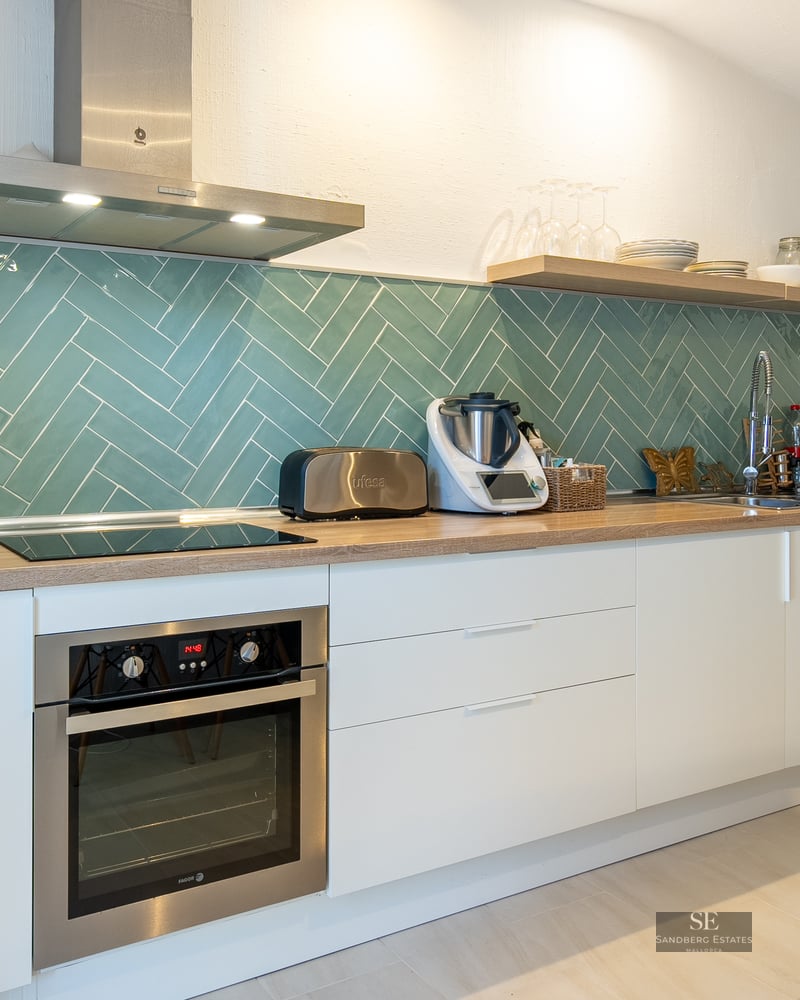 Modern white kitchen featuring a teal herringbone tile backsplash, wooden countertops, and stainless steel appliances.