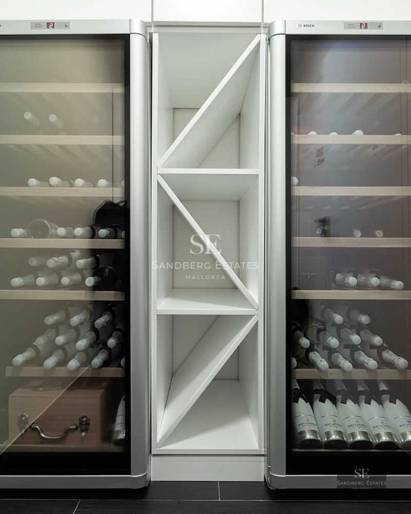 Two tall glass-front wine coolers with wooden shelves flanking a white geometric storage unit on a dark tile floor.
