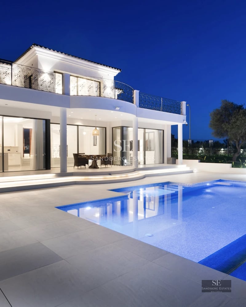 View of reflective infinity pool, modern architecture, large windows, and lush vegetation. Soft ambient lighting.