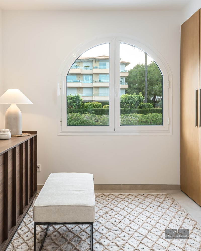 A modern dressing room featuring large oak wardrobes, an arched window, a mirrored console, and a cushioned bench on a rug.