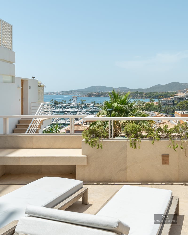 Sun loungers on a modern stone terrace overlooking a marina with boats and mountains under a clear blue sky.