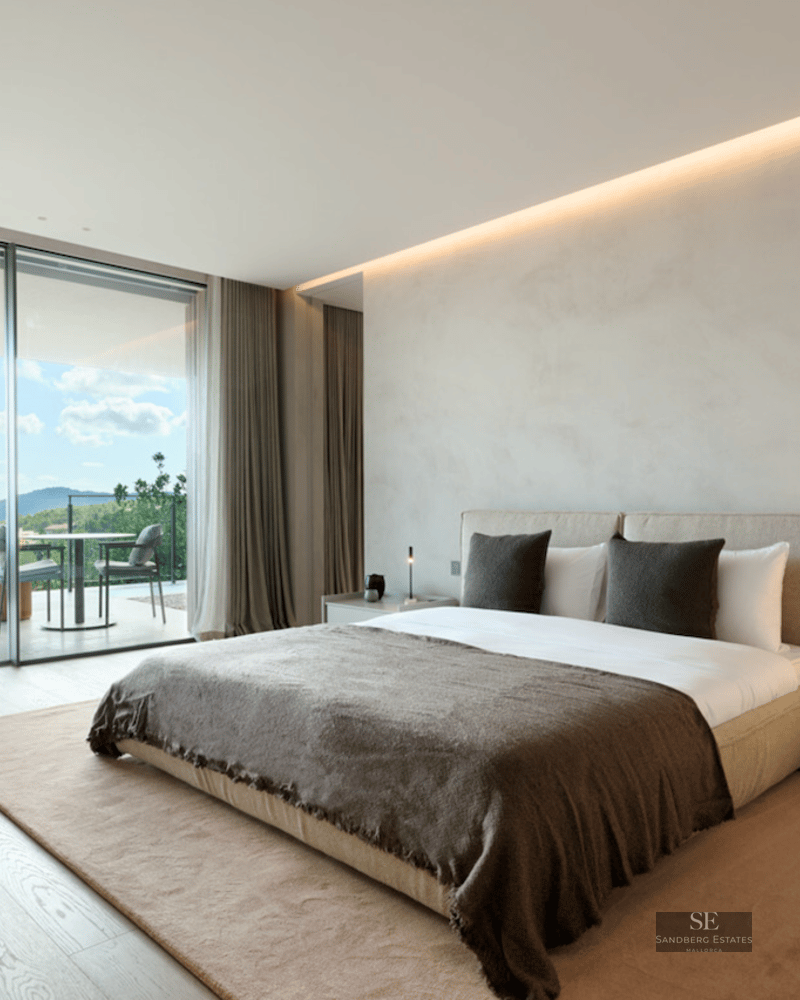 Modern master bedroom with a large bed, designer armchair, and floor-to-ceiling glass doors opening to a scenic terrace.