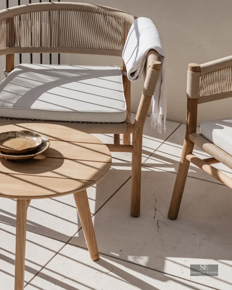 Two elegant woven chairs and a round wooden side table on a sunlit balcony with shadows.