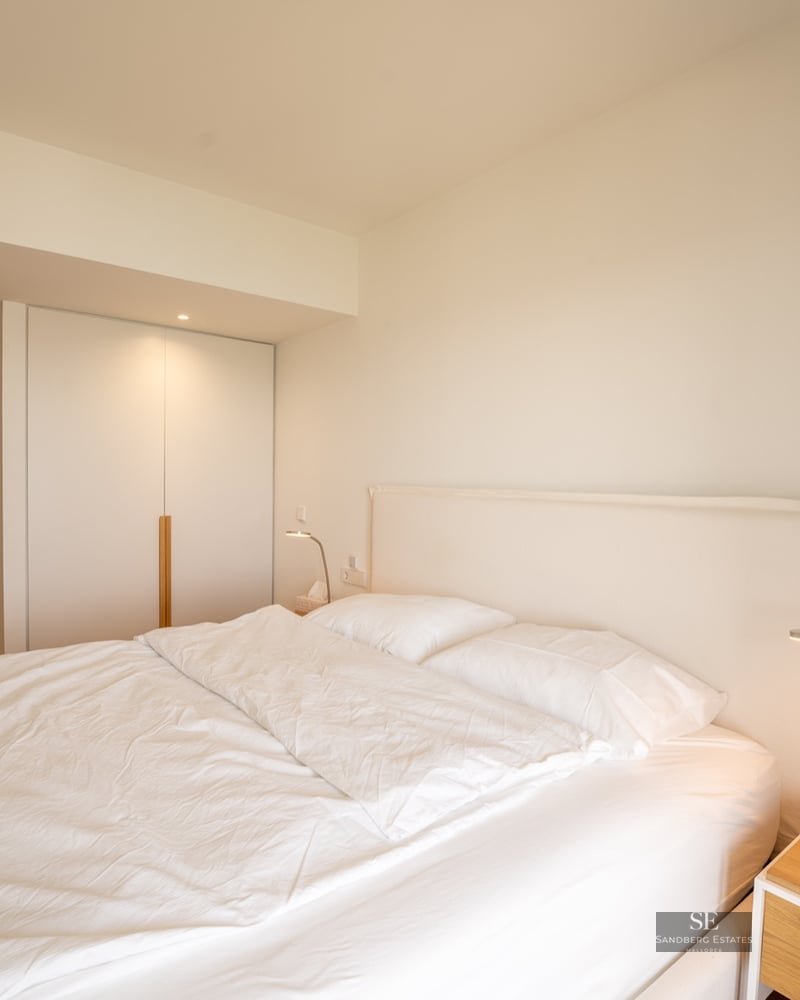 Bright minimalist bedroom with a large bed, white linens, built-in closet, and wooden bedside table.