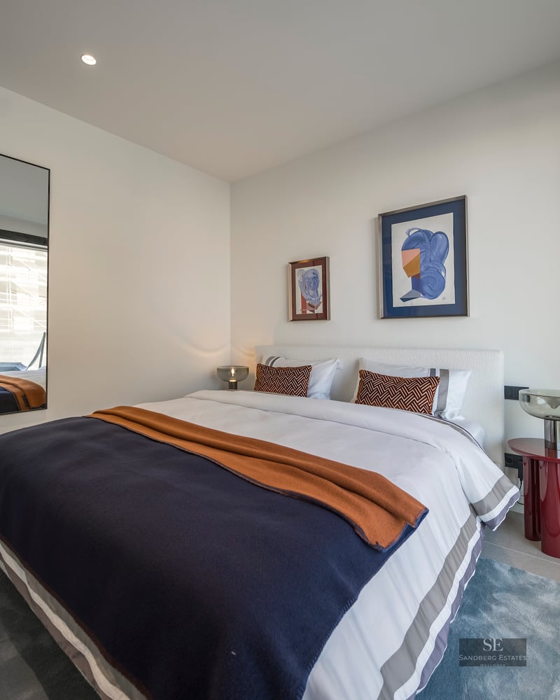 A bright modern bedroom with a large bed, navy and orange bedding, a red side table, and a large mirror reflecting a window.