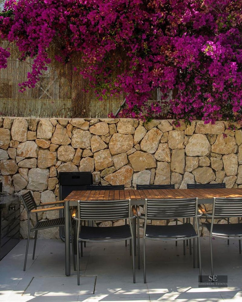 Outdoor patio featuring a wooden dining table, black chairs, gas grill, and a stone wall with vibrant pink flowers.