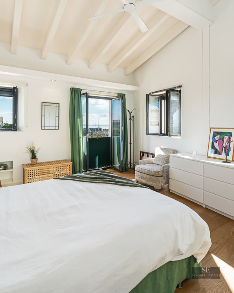 Bright bedroom with white walls, wooden beams, white bed, and windows overlooking a harbor and cathedral.