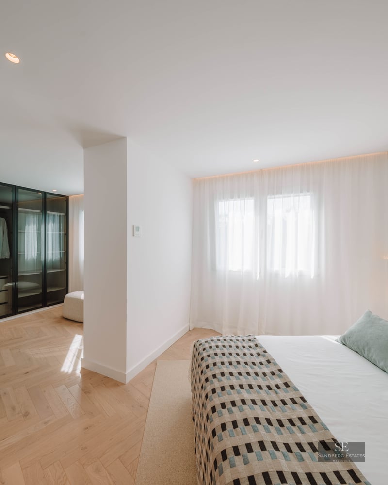 Bright master bedroom featuring herringbone wood floors, a large bed with pillows, and a glass-front walk-in closet.