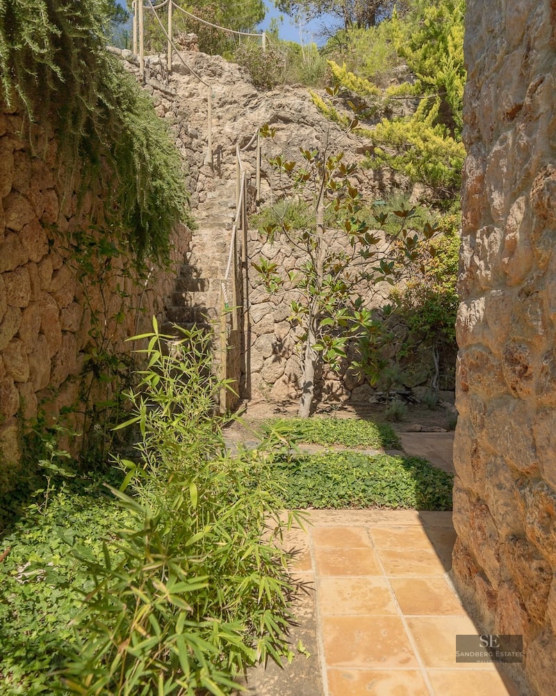 Terracotta tile path between rustic stone walls and lush greenery leading to stone stairs.