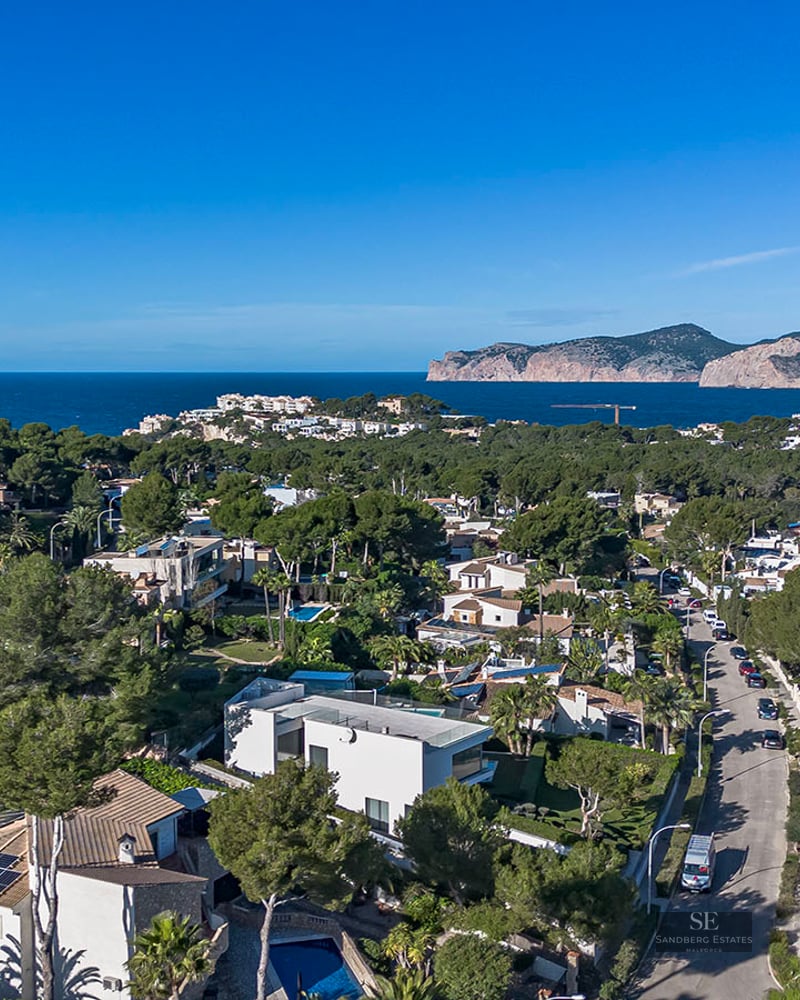 Drone shot of a coastal neighborhood featuring luxury homes, pine trees, and the Mediterranean Sea in the background.