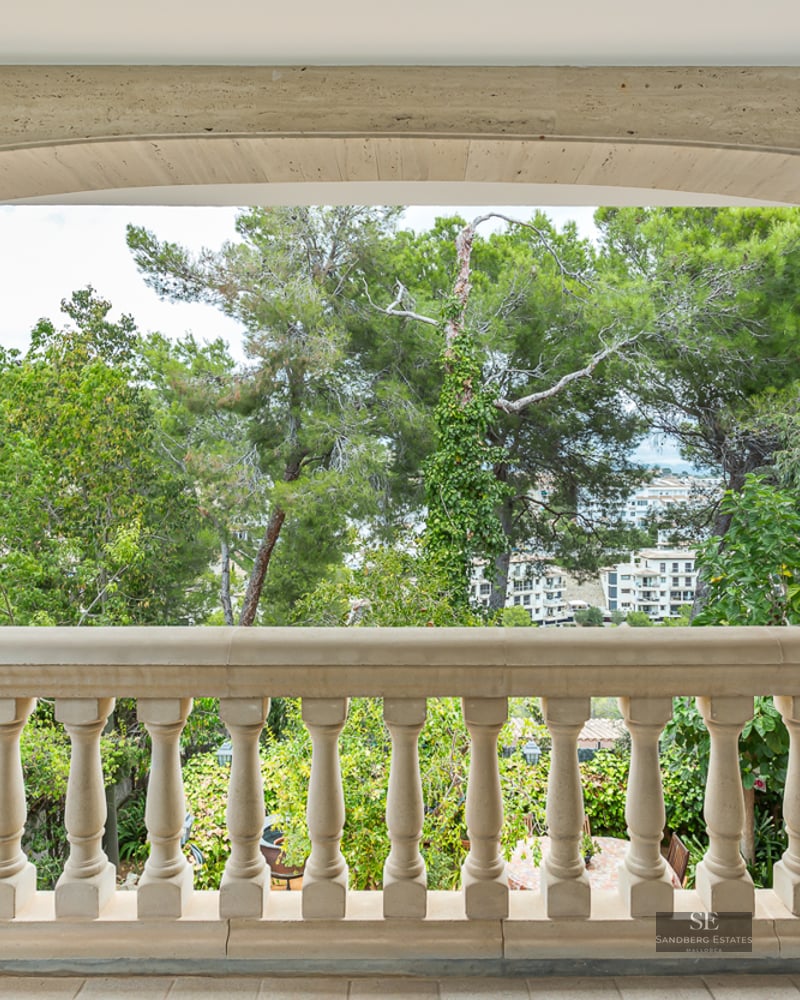 A beige stone balustrade under a wide stone arch overlooking lush green trees and distant buildings.