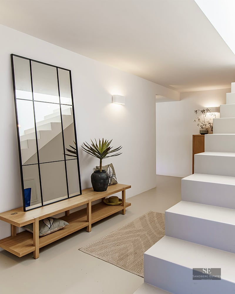 Bright minimalist hallway with white stairs, a large black-framed mirror, wooden bench, and warm wall lighting.