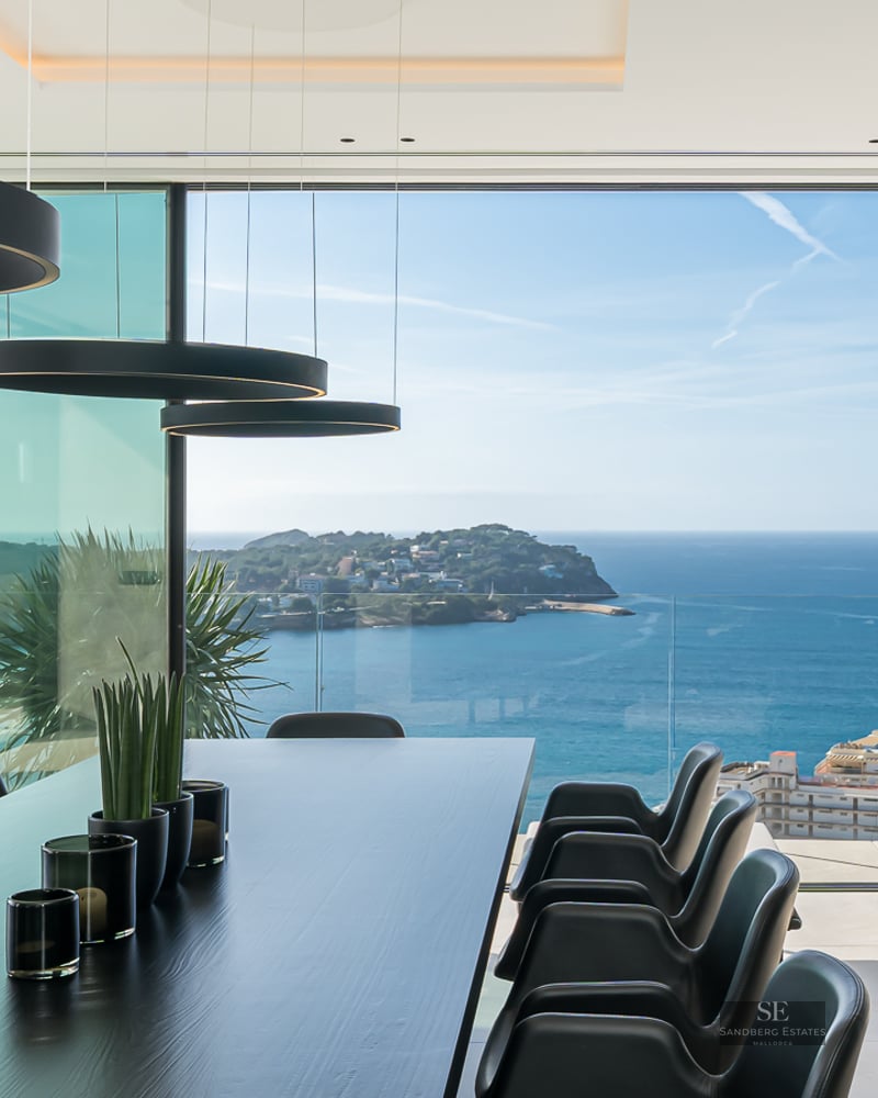 Modern black dining table and chairs facing floor-to-ceiling glass walls overlooking the blue ocean and coastline.