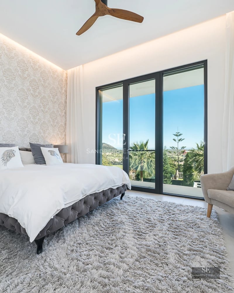 Panoramic view from a balcony with glass railing overlooking the sea. Modern and bright interior. Potted plants.