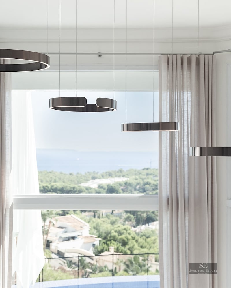 Four bronze ring-shaped pendant lights hanging in front of a large bay window overlooking a coastal landscape and the sea.
