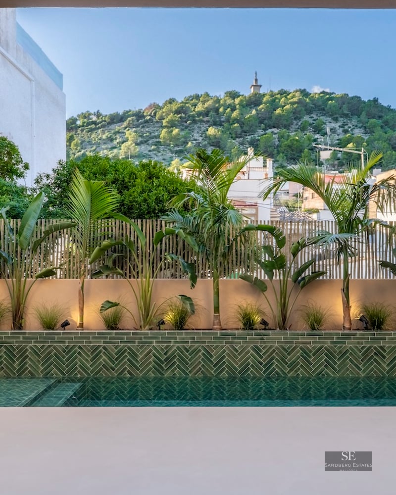Piscine moderne avec carreaux verts en chevrons entourée de plantes tropicales et vue sur une colline sous un ciel bleu.