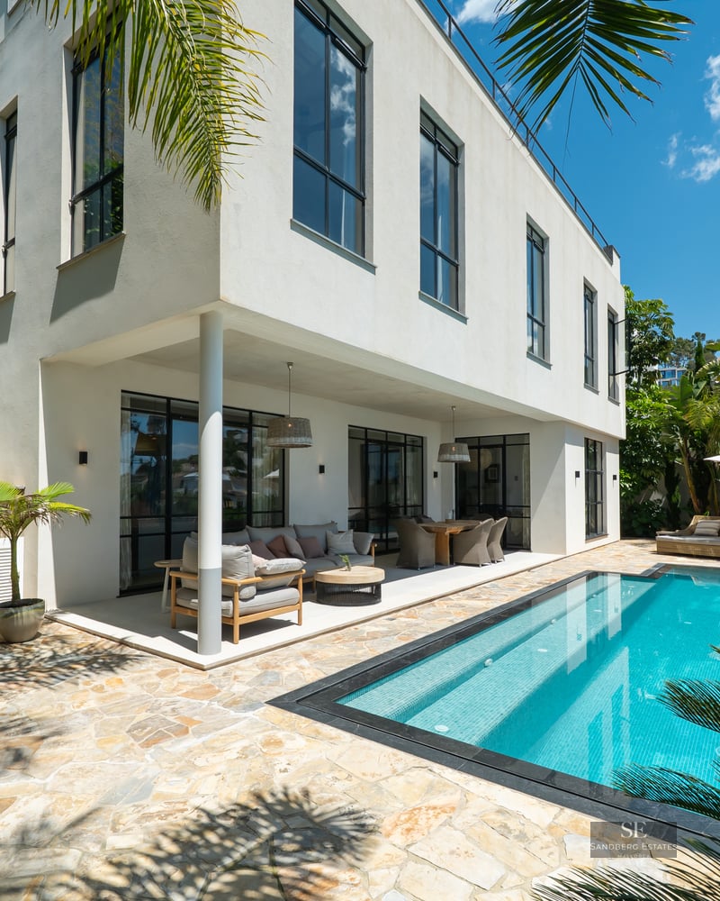 A bright outdoor scene featuring a clear turquoise swimming pool, stone-paved terrace, and a modern white villa.