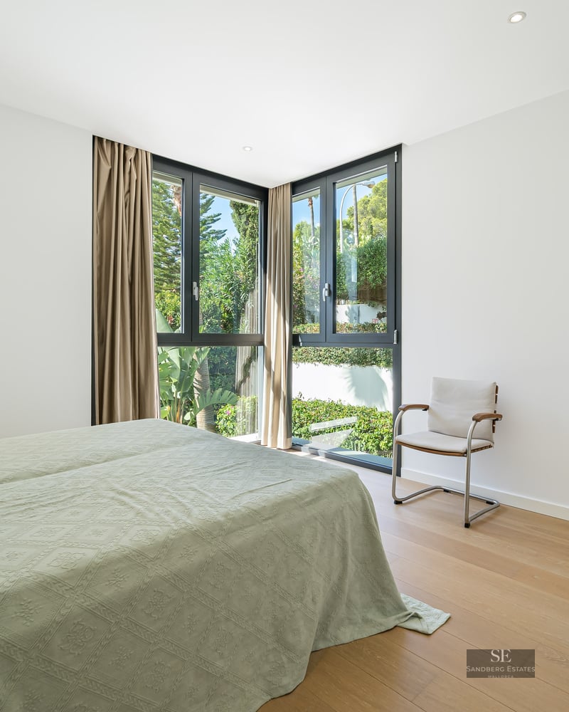 Bright master bedroom with wood floors, large corner windows overlooking greenery, and an adjacent modern bathroom.