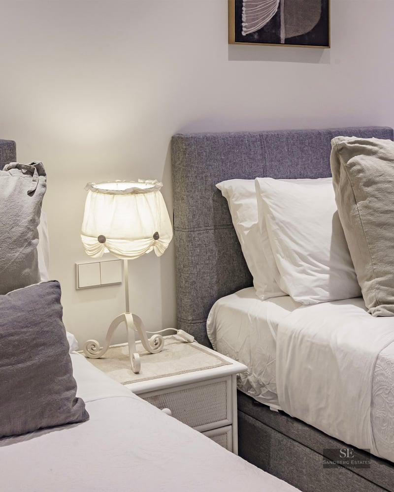 Two twin beds with grey upholstered headboards, white linens, and a lamp on a bedside table between them.