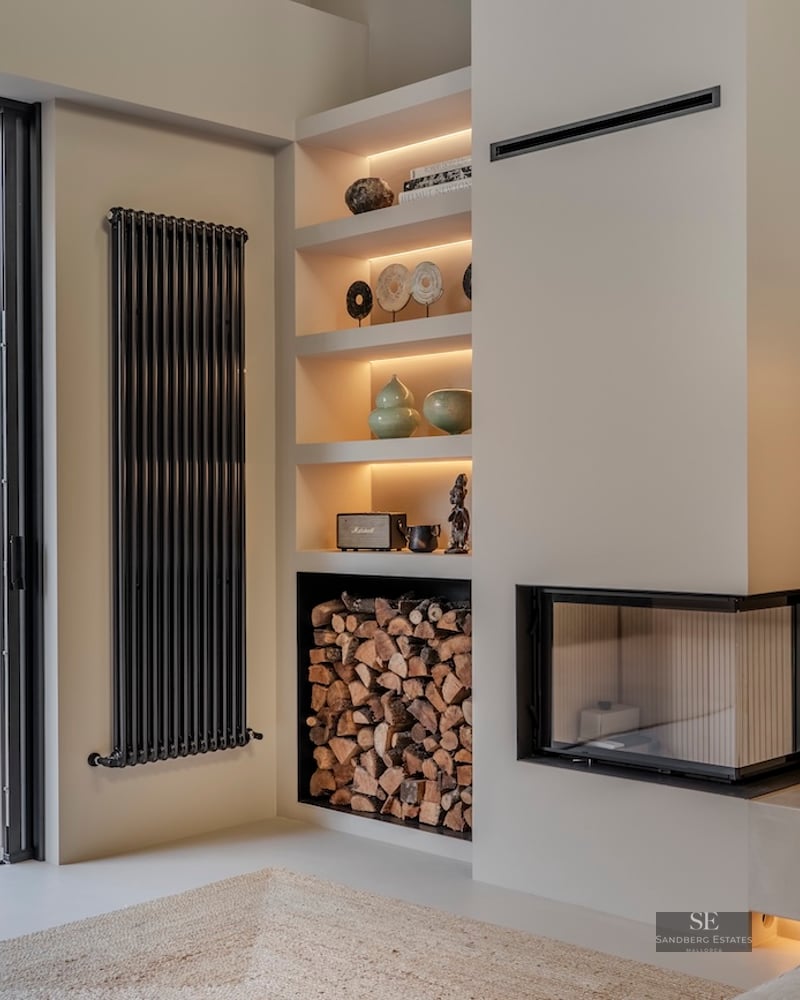 Espace de vie intérieur avec cheminée d'angle moderne, rangement à bois et vue sur une terrasse extérieure luxuriante.