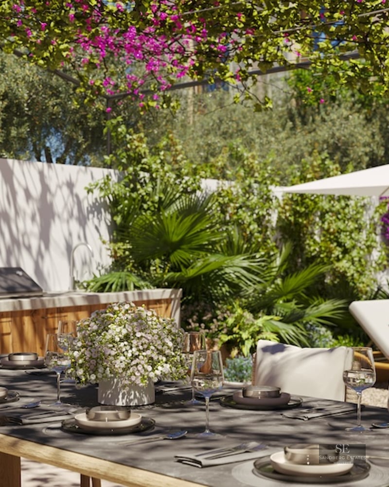 An outdoor dining table set under a bougainvillea pergola, featuring an outdoor kitchen and sun loungers in a garden.