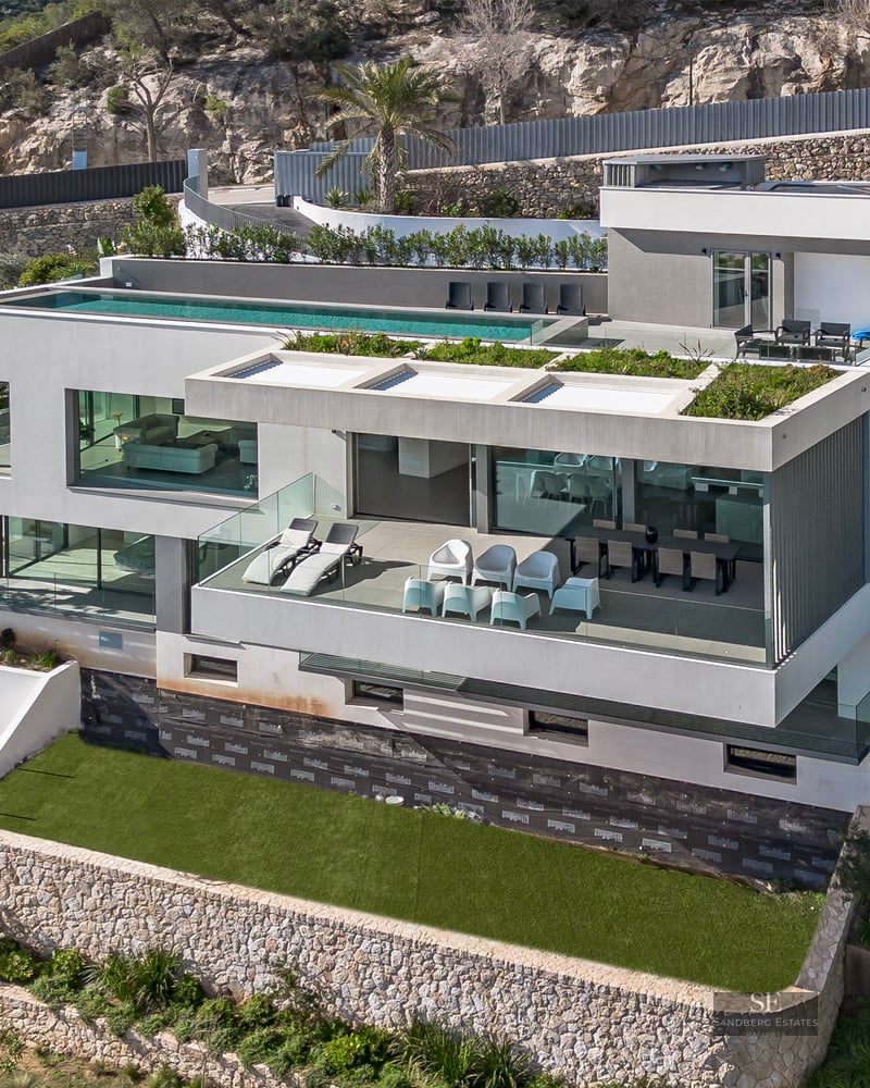 Aerial view of a white multi-story modern villa with a rooftop infinity pool, glass terraces, and stone walls on a hill.