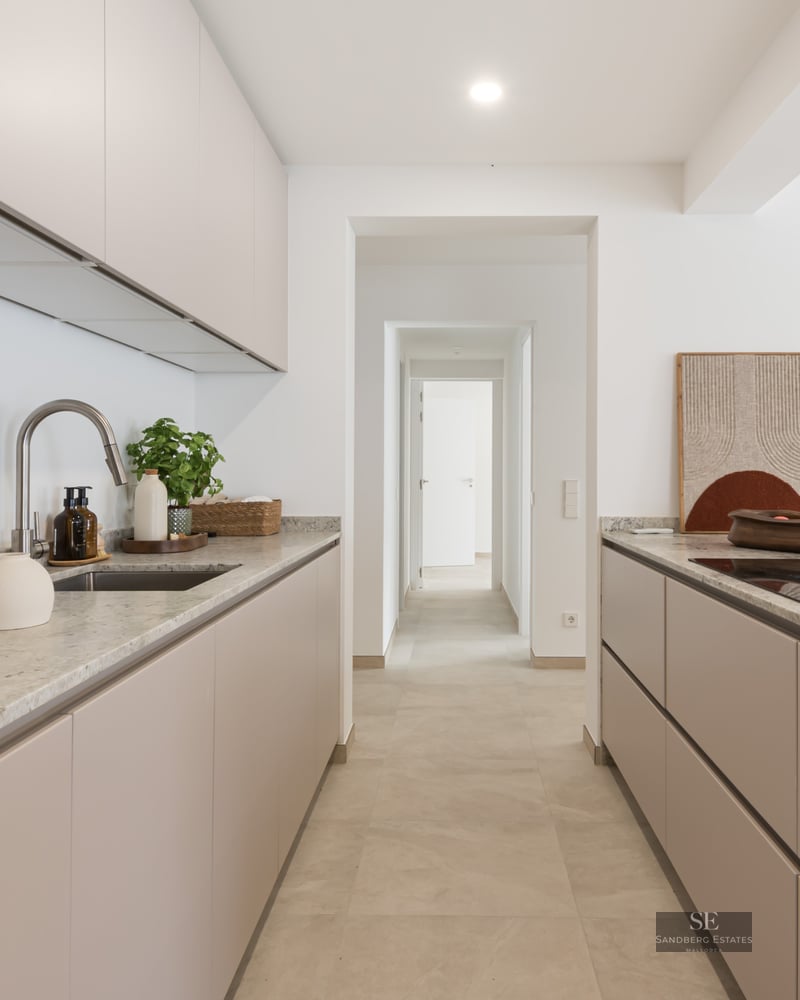 Modern kitchen with beige cabinets, stone countertops, and a view down a white hallway.