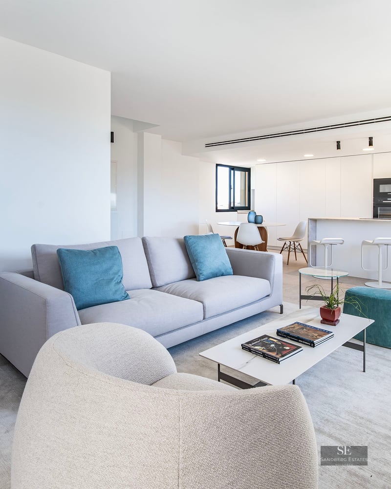 Modern living room with grey sofa, teal pillows, marble coffee table, and spiral staircase next to a white kitchen.