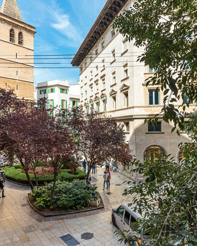 Pedestrian plaza with trees, stone buildings, and a church tower under a clear blue sky.
