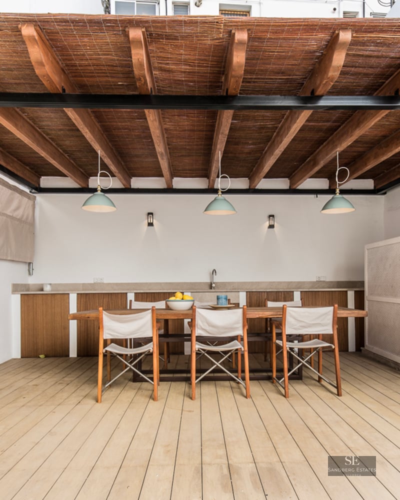 Shaded outdoor dining area with a large wooden table, chairs, and a pergola with integrated lighting and kitchenette.