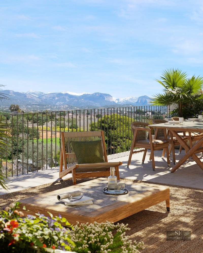 Sunny terrace with wooden furniture and rug overlooking a valley and snow-capped mountains.