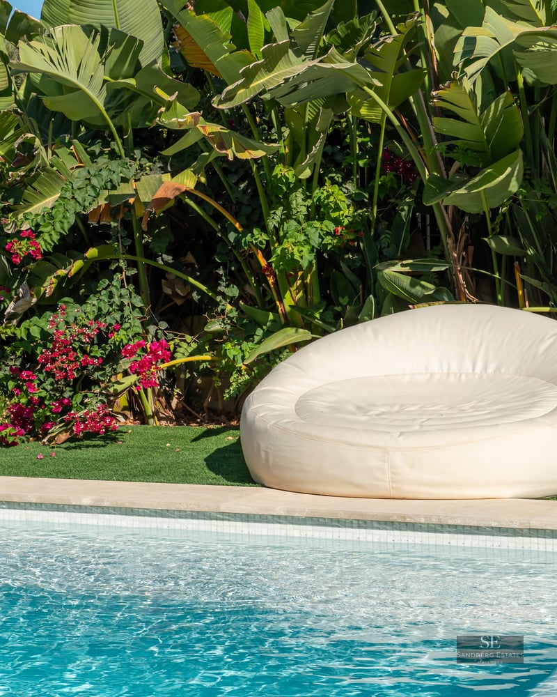 A white circular outdoor lounger beside a blue swimming pool, surrounded by lush tropical plants and pink flowers.