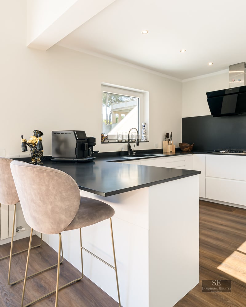 Cuisine moderne blanche avec plans de travail noirs, parquet et bar donnant sur une terrasse ensoleillée.