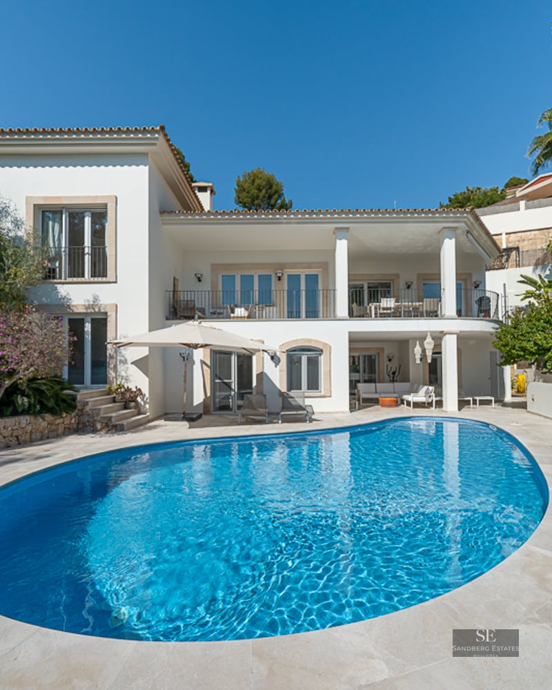 Grande piscine ovale bleu azur devant une villa blanche à deux étages avec terrasse en pierre et palmiers.