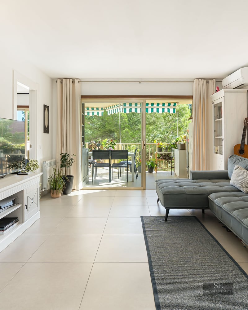 Salon moderne avec canapé bleu, meuble TV blanc et baies vitrées ouvrant sur une terrasse verdoyante.