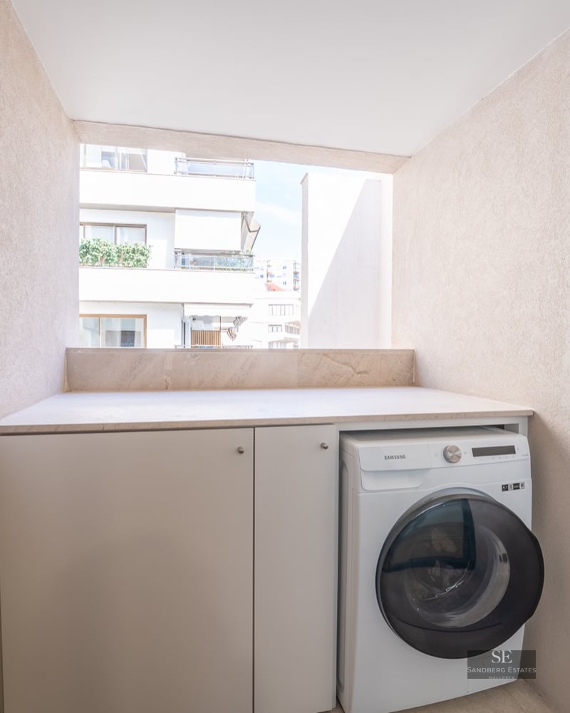 Modern washing machine and white cabinets in a textured beige balcony nook overlooking city buildings.
