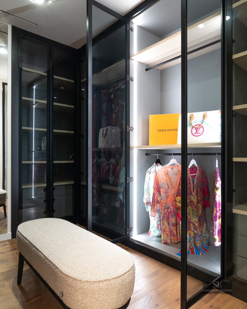 Modern walk-in closet featuring glass-door wardrobes with LED lighting, wood floors, and a textured boucle bench.