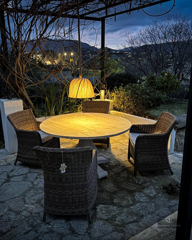 Outdoor stone terrace with a round wooden table, wicker chairs, and a warm pendant light overlooking hills at dusk.