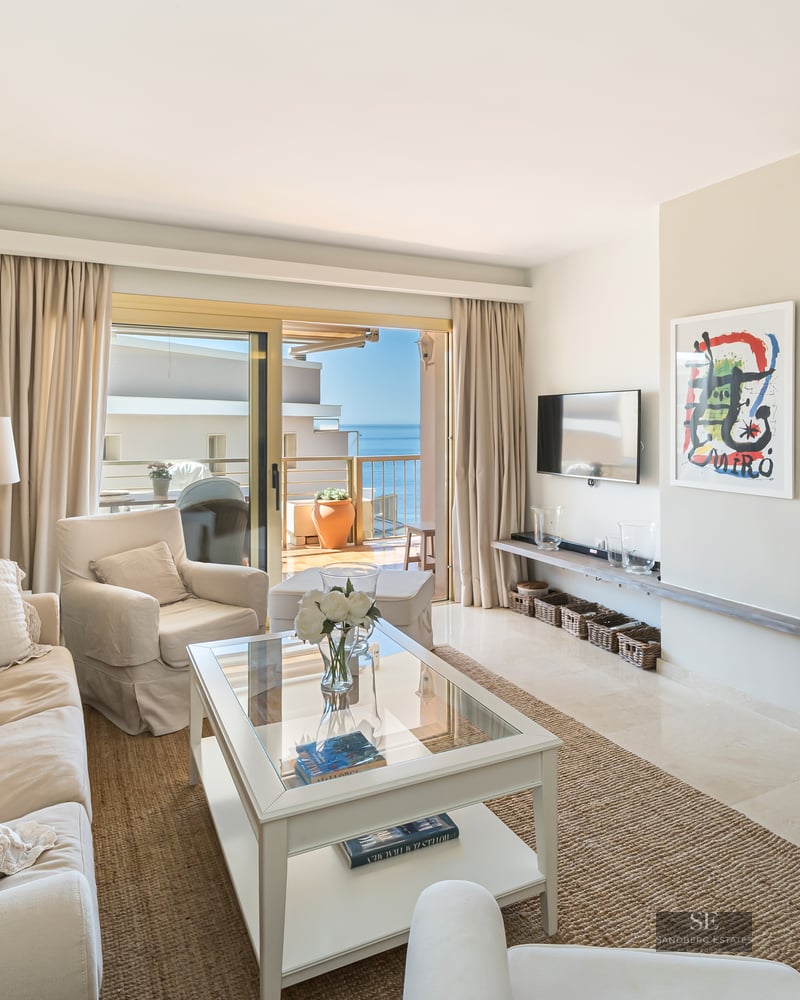 Mediterranean style living room with beige sofas, glass table, and sliding doors opening to a terrace with sea views.