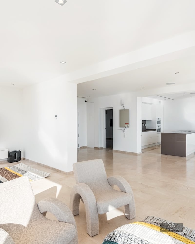 Bright, modern living room with plush armchairs, a large wall-mounted TV, and an adjacent open-plan kitchen.