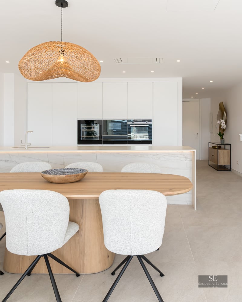 A bright dining area with a wooden table, white chairs, and a wicker pendant light in front of a modern kitchen.