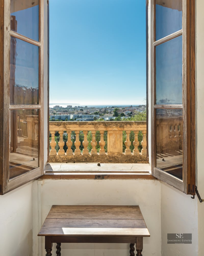 An open wooden window framing a panoramic view of a coastal city and the sea under a clear blue sky.