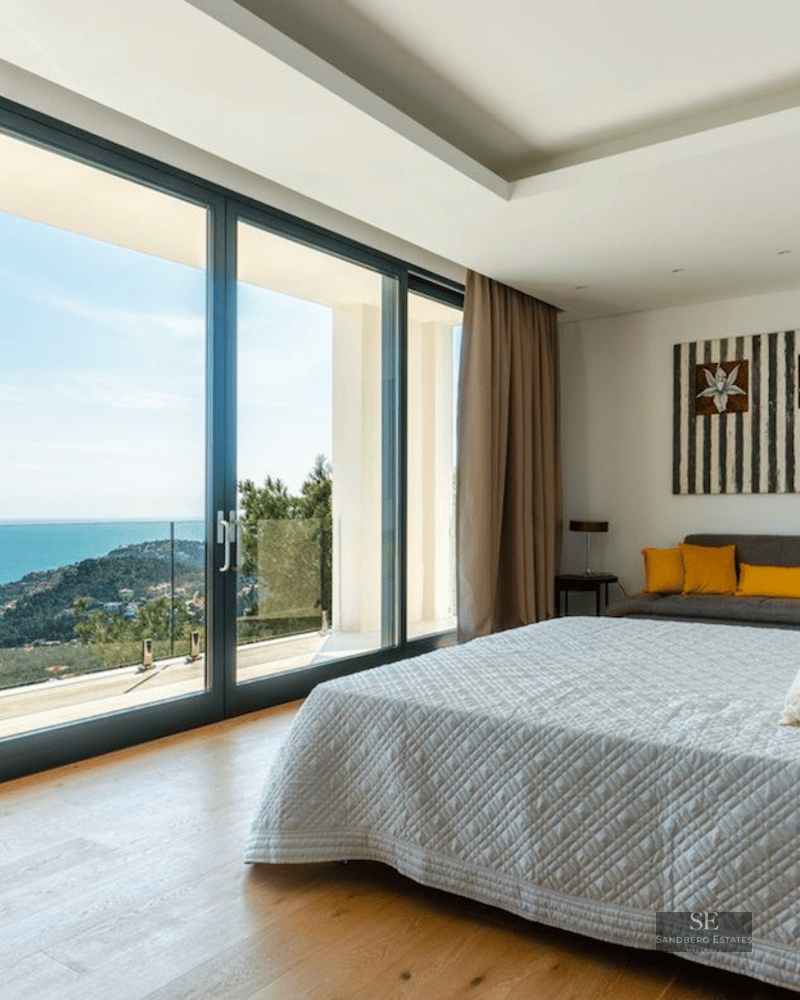 Modern master bedroom featuring floor-to-ceiling glass doors with a stunning view of the sea and coastline.