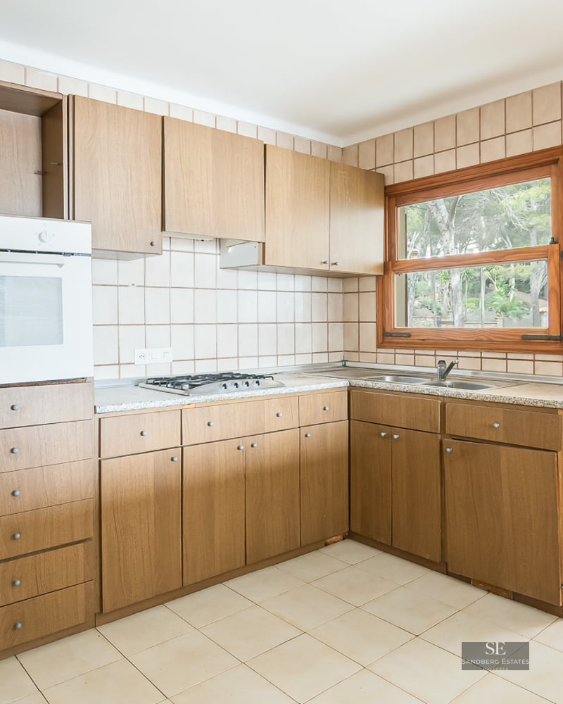 Bright kitchen featuring wooden cabinetry, beige wall tiles, and views of greenery through a window and door.