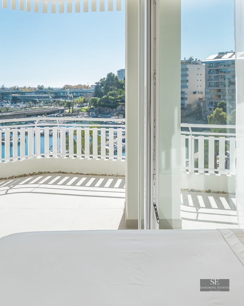 Bright bedroom with white bed and open glass doors leading to a balcony overlooking a marina with boats.